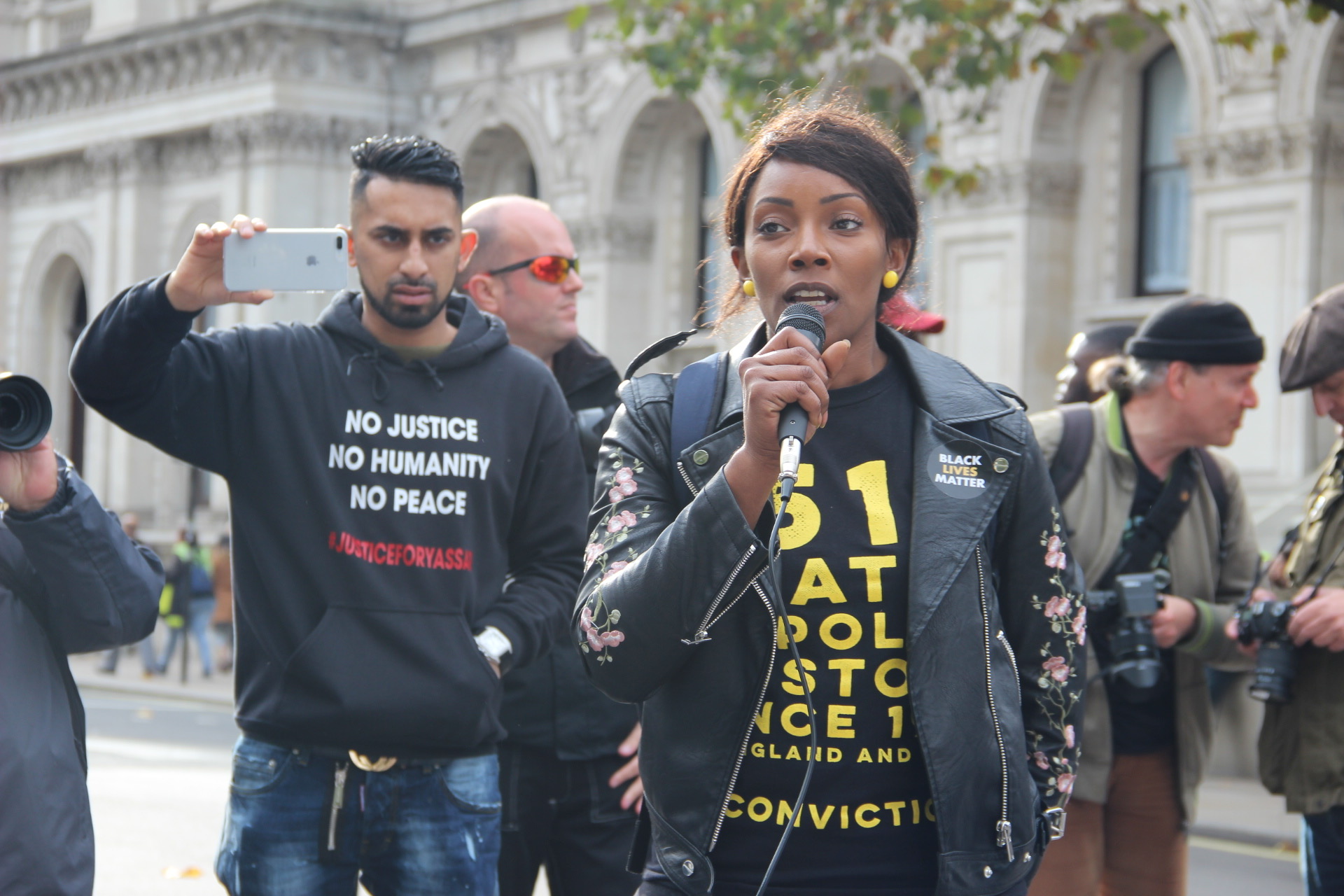 Woman making a speech on a microphone while an onlooker films her using his cameraphone