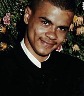 A photo of Mark Duggan. He is smiling at the camera, wearing a white t shirt with a black garment on top, and there are flowers in the background.