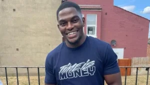 A photograph of Chris Kaba. He is outside in front of a house, smiling at the camera and wearing a t shirt that says 'Time Is Money'.